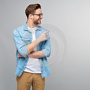 Happy young handsome man in jeans shirt pointing away standing against grey background