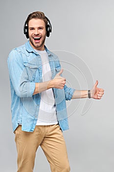 Happy young handsome man in jeans shirt pointing away standing against grey background wearing big headphones