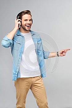 Happy young handsome man in jeans shirt pointing away standing against grey background wearing big headphones