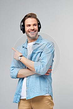 Happy young handsome man in jeans shirt pointing away standing against grey background wearing big headphones