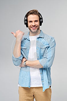 Happy young handsome man in jeans shirt pointing away standing against grey background wearing big headphones