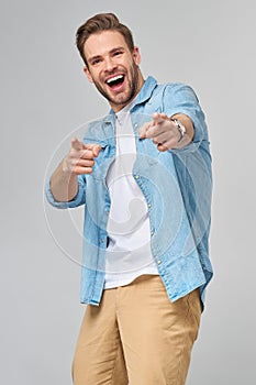 Happy young handsome man in jeans shirt pointing on camera standing against grey background