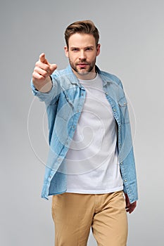 Happy young handsome man in jeans shirt pointing on camera standing against grey background