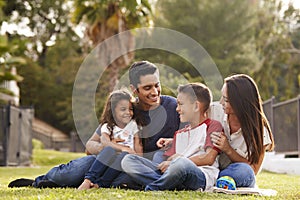 Happy young Hispanic family sitting together on the grass in the park, looking at each other