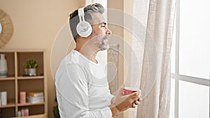 Happy young hispanic man, grey-haired and handsome, casually drinking coffee and enjoying music at home in the morning, listening