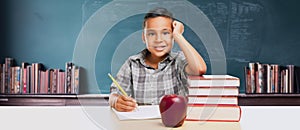 Happy Young Hispanic School Boy At Desk with Books and Apple In Front of Chalkboard