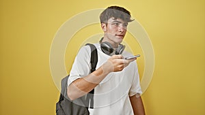 Happy young hispanic student guy sending a sweet voice message on his smart phone, standing against isolated yellow wall with