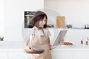 Happy young housewife cooking in kitchen, wearing apron and browsing recipes via digital tablet, holding frying pan