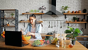 Happy young housewife preparing vegetable salad in the kitchen while looking at a recipe on the internet on her computer