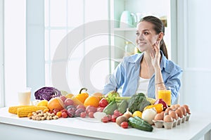 Happy young housewife sitting in the kitchen preparing food from a pile of diverse fresh organic fruits and vegetables