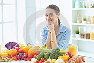 Happy young housewife sitting in the kitchen preparing food from a pile of diverse fresh organic fruits and vegetables