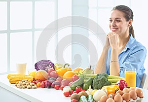 Happy young housewife sitting in the kitchen preparing food from a pile of diverse fresh organic fruits and vegetables