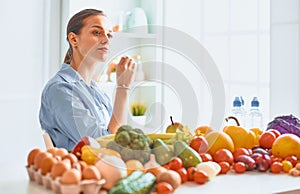 Happy young housewife sitting in the kitchen preparing food from a pile of diverse fresh organic fruits and vegetables