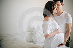 Happy young husband kissing his smiling pregnant wife and hugging belly bump on white bed. Stylish pregnant couple in white