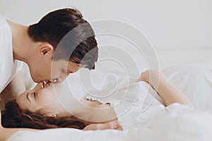 Happy young husband kissing his smiling pregnant wife and hugging belly bump on white bed. Stylish pregnant couple in white