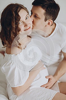 Happy young husband kissing his smiling pregnant wife and hugging belly bump on white bed. Stylish pregnant couple in white