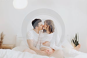 Happy young husband kissing his smiling pregnant wife and hugging belly bump on white bed. Stylish pregnant couple in white