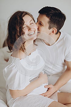 Happy young husband kissing his smiling pregnant wife and hugging belly bump on white bed. Stylish pregnant couple in white