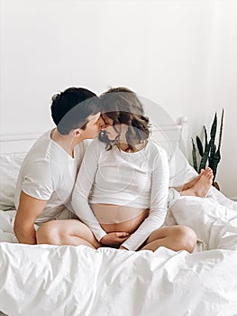 Happy young husband kissing his smiling pregnant wife and hugging belly bump on white bed. Stylish pregnant couple in white