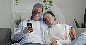 Happy young husband showing funny video in social network on cellphone beautiful wife. Smiling afro american spouses