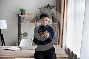 Happy young Indian businesswoman using cellphone leaning on table.