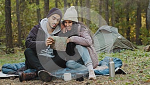 Happy young indian couple hikers sit in nature near tent look at paper map check route directions using guidebook two