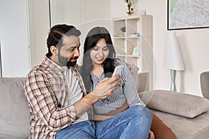 Happy young indian couple using phone sitting on sofa together at home.