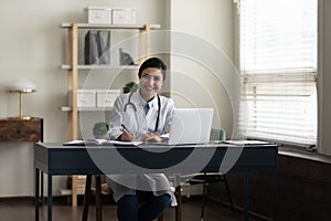 Happy young Indian doctor, therapist, general practitioner at work table