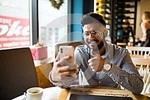 Happy young indian man sitting at cafe making video call from his mobile phone. Asian male at coffee shop having a videochat on