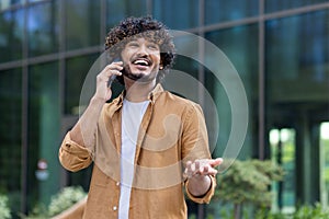 A happy young Indian man is standing on a city street and talking emotionally on a mobile phone