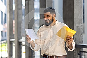 Happy young Indian man standing outside building, holding envelope and smilingly reading received letter and results photo