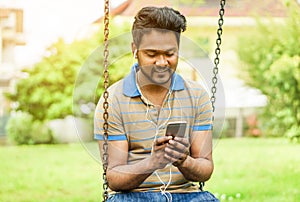 Happy young Indian man watching videos and sharing with mobile cell phone - University student using smartphone outdoor - New