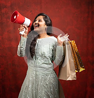 Happy Young indian woman making annnouncement on megaphone about online or e-commerce sales by holding shopping bags - concept of
