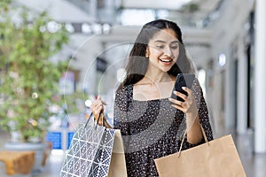 A happy young Indian woman is shopping in a shopping center, standing inside, holding paper bags with goods and using a