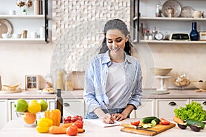 Happy young lady making notes on table with organic vegetables, cooking dinner in light kitchen interior