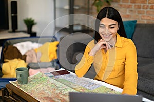 Happy young lady planning vacation with map and laptop, checking best travel routes for journey, preparing for trip