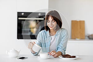 Happy young lady pouring milk in cup of coffee, sitting in kitchen interior, enjoying morning hot beverage