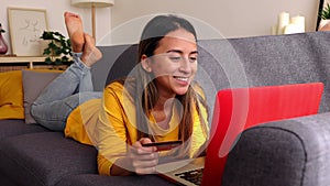 Happy young Latin American woman doing online shopping lying on sofa at home.