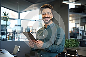 Happy young Latin business man holding tablet standing in office. Portrait