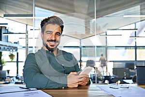 Happy young Latin business man using mobile phone working in office, portrait.
