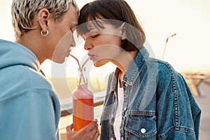 Happy young lesbian couple drinking from one glass bottle with the straw, Two women enjoying cold beverage on a summer