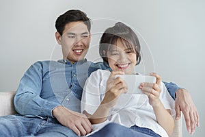Happy young love asian couple sitting on couch at home, looking at mobile phone, Asian teen couples are enjoying the game
