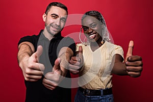 Happy young lovely interracial couple showing thumbs up and looking at the camera over red background