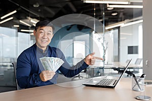 Happy young man Asian businessman sitting in the office at the table holding cash money in his hands