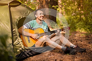Happy young man camping and strum a guitar instrumental music to relax against background of forest sunset.
