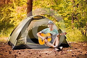 Happy young man camping and strum a guitar instrumental music to relax against background of forest sunset.