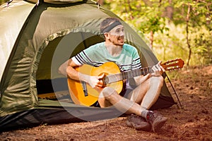 Happy young man camping and strum a guitar instrumental music to relax against background of forest sunset.