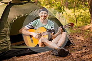 Happy young man camping and strum a guitar instrumental music to relax against background of forest sunset