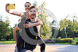 Happy young man carrying his girlfriend on his back after training in park and she taking selfie