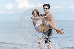 Happy young man carrying woman with arm up and walks on beach in wedding vacation, honeymoon couple are both laughing and funny